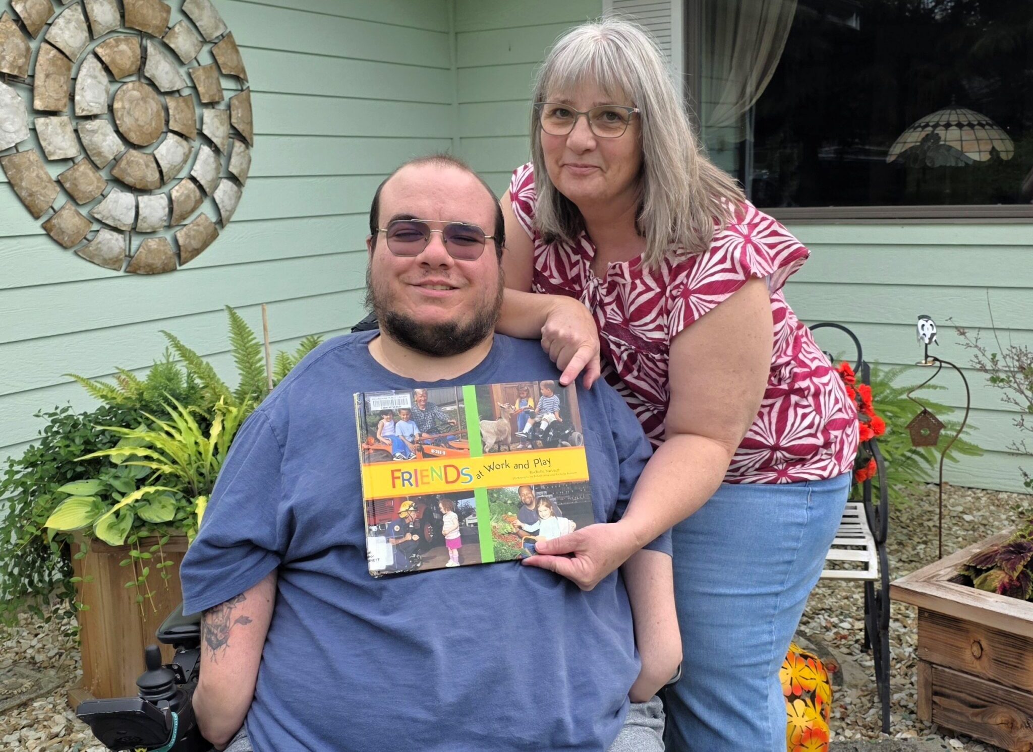 An adult man in a wheelchair and his mother holding up a book and pointing to a picture of him as a child on the cover.