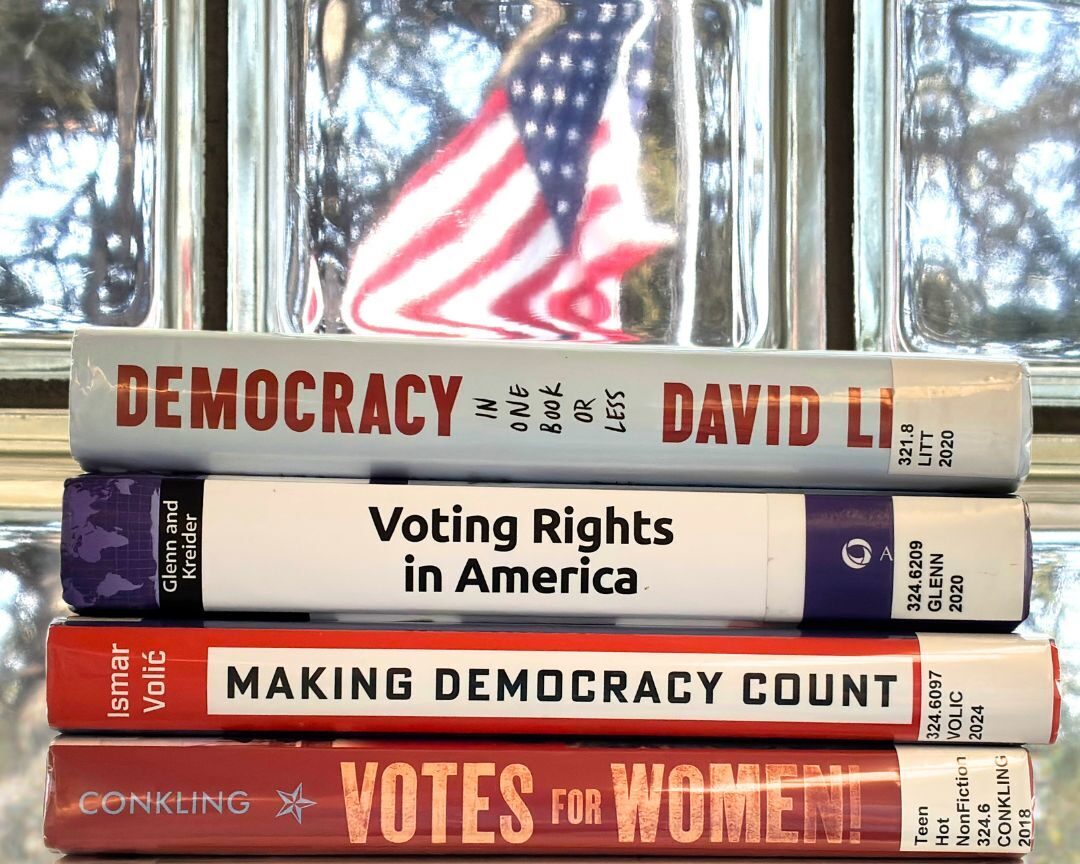 A stack of books about democracy in front of a window with an American flag in the background